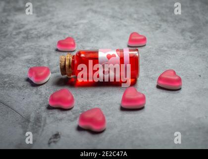 Image en gros plan d'une potion d'amour rouge dans un verre bouteille de bouchon entourée de bonbons en forme de cœur rose une surface texturée en béton Banque D'Images