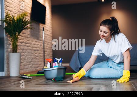 Jolie femme en t-shirt blanc avec cheveux foncés de nettoyage dans des gants en caoutchouc jaune pour la protection des mains et seau avec des fournitures de nettoyage à la maison. Banque D'Images