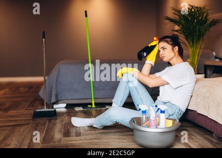 Femme fatiguée en t-shirt blanc avec cheveux foncés nettoyage dans des gants en caoutchouc jaune pour la protection des mains et seau avec des fournitures de nettoyage à la maison. Banque D'Images
