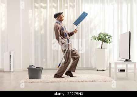 Homme âgé chantant et dansant avec une mop de nettoyage à maison devant la télévision dans un salon Banque D'Images