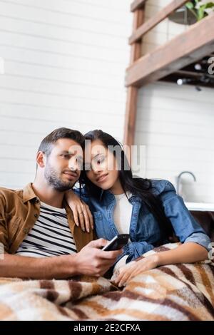 une femme heureuse avec des yeux fermés penchée sur l'épaule d'un petit ami regardant la télévision sous une couverture à carreaux, avant-plan flou Banque D'Images