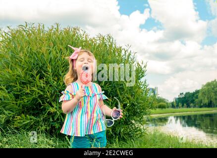 Jeune fille à la mode, lunettes de soleil, lipop comme un coeur.petite fille de trois ans lèche un bonbon tourbillonnant coloré lors d'une promenade dans le parc le jour d'été Banque D'Images