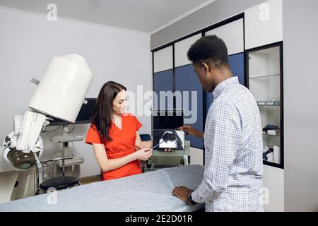 Jeune femme médecin caucasien dans une clinique moderne, tenant un PC tablette et montrant à son patient africain d'échographie des reins et interne Banque D'Images