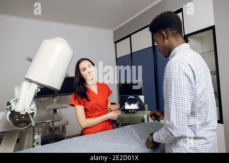 Jeune femme médecin caucasien dans une clinique moderne, tenant un PC tablette et montrant à son patient africain d'échographie des reins et interne Banque D'Images