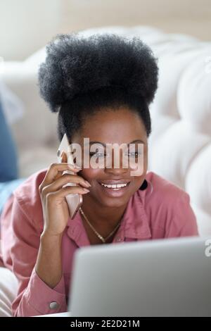 Bonne femme africaine couchée sur un canapé à la maison ordinateur portable parlant sur un smartphone mobile Banque D'Images