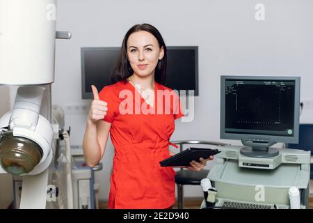 Gros plan de la jolie femme caucasienne médecin debout devant les échographes et les appareils de lithotripter et montrant le pouce vers le haut. Pierre non invasive de sécurité Banque D'Images