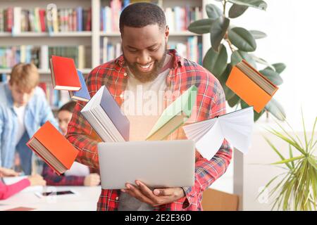 Étudiant afro-américain avec livre de lecture en ligne d'ordinateur portable dans la bibliothèque Banque D'Images