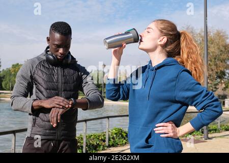 Homme noir africain et femme caucasienne buvant de l'eau et mettant en place une montre intelligente. Fitness et sport en plein air. Banque D'Images