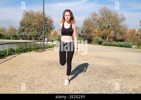 REDHEAD femme caucasienne avec des écouteurs qui étirent sa jambe. Fitness et sport en plein air. Banque D'Images