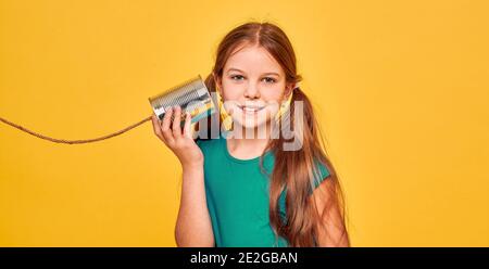 Adolescente souriante écoutant de l'étain CAN téléphone, jouant dans le chat rétro connecté, sur fond jaune. Concept de câbles croisés et de connexion téléphonique Banque D'Images