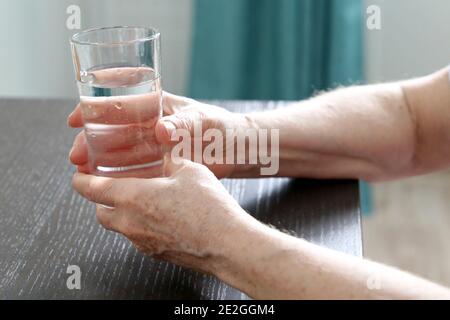 Femme âgée avec un verre d'eau propre dans les mains froissées à la table. Concept de soif, régime à la retraite, purification de l'eau, boisson minérale Banque D'Images