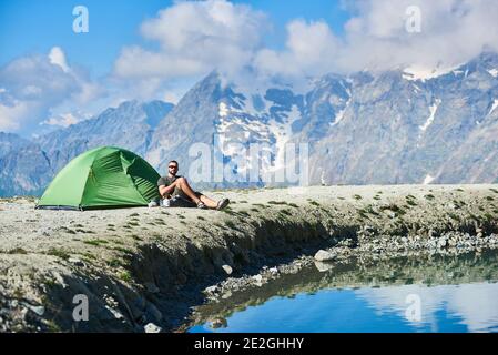 Homme voyageur assis près d'une tente touristique près d'un beau lac bleu avec de l'eau cristalline. Randonneur se reposer dans les montagnes avec de majestueux pics brumeux en arrière-plan. Concept de voyage, de randonnée et de camping. Banque D'Images