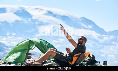 Homme touriste assis dans une chaise près d'une tente dans des montagnes rocheuses vides, en utilisant un smartphone dans une chaude journée d'été ensoleillée et pointant avec la main vers le haut. Concept de camping, voyage dans les Alpes, nature, gadgets. Banque D'Images