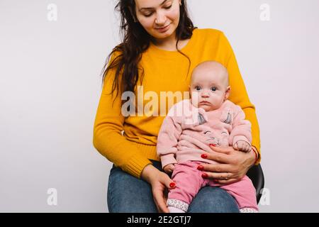 Une petite fille avec de grands yeux bleus regarde la caméra tout en étant assise dans les bras de sa mère après avoir percé l'oreille sur fond gris. Banque D'Images