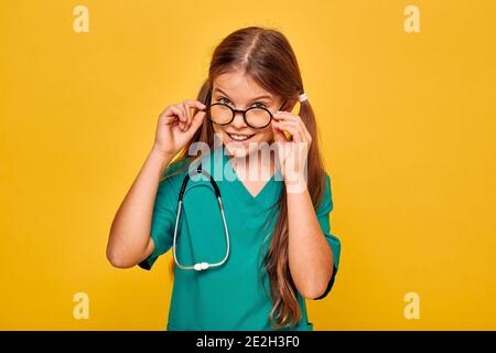 Fille portant un uniforme chirurgical et un stéthoscope jouant la profession médicale, et montre sur son visage l'émotion de surprise, en prenant des lunettes, sur vous Banque D'Images