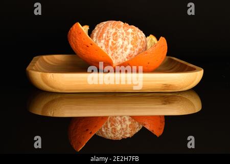 Une mandarine mûre juteuse orange vif entière, à moitié pelée, se trouve sur un plateau en bambou, sur fond noir. Banque D'Images