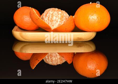 Deux mandarines entières et une mandarine mûre juteuse orange vif, à moitié pelée, se trouvent sur un plateau en bambou, sur fond noir. Banque D'Images