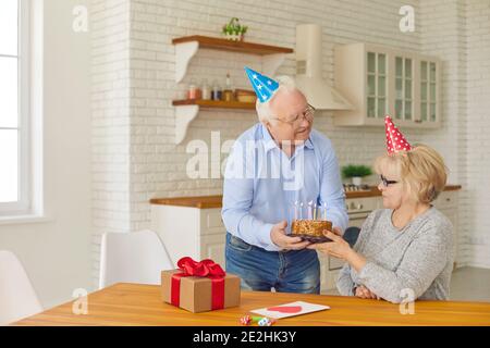 Romantique homme plus âgé donne un gâteau à sa femme pour faire un souhait et souffler les bougies. Couple senior en chapeaux coniques de couleur célébrant l'anniversaire ou Banque D'Images