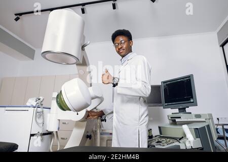 Portrait d'un jeune homme africain professionnel, un médecin debout près des échographes et des lithotripteurs modernes et montrant le pouce vers le haut. Sécurité non Banque D'Images