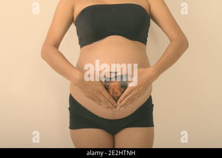 Studio shot of young pregnant woman holding sonogramme de son bébé tout en faisant signe à main coeur contre fond blanc Banque D'Images