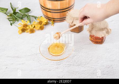 La main d'une femme tient une cuillère en bois sur une soucoupe de miel, un baril en céramique et des pots de miel sur un fond clair. Banque D'Images