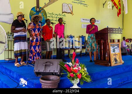 Plusieurs chanteuses et un chanteur se tiennent avec des micros sur la scène d'une église baptiste à The Lime, Grenade, tandis qu'une femme en blazer rose parle à la tribune. Une fresque murale peinte représentant une croix, un globe et une citation biblique décore le mur derrière eux, avec un drapeau grenadien accroché à droite. Banque D'Images