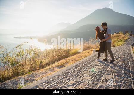 Un couple de voyageurs s'embrassant au milieu d'une route à Lever du soleil avec le lac Atitlan derrière eux - couple hispanique dedans amour avec les volcans et les montagnes Banque D'Images