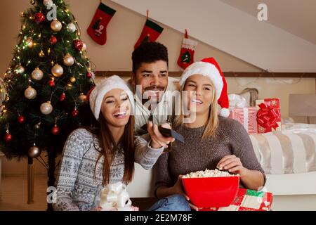 Amis regardant la télévision et mangeant du pop-corn tout en étant assis dans la chambre. Concept des fêtes de Noël. Banque D'Images