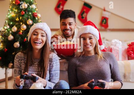 Deux amies jouant à des jeux vidéo en étant assise sur le sol. Leur ami allongé sur le lit avec du pop-corn entre les mains et les regardant. Noël en arrière-plan Banque D'Images