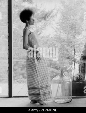 ANNÉES 1970 SLIM AFRO-AMÉRICAINE FEMME AFRO COIFFURE PIEDS NUS DANS LA MODE ROBE LONGUE AVEC CORSAGE SMOCKÉ AVANT DE TENIR UNE FENÊTRE - G8170 HAR001 HARS COIFFURE NOIR ETHNICITÉ PIEDS NUS POSANT BODICE ÉLÉGANT LA MODÉLISATION DES MODES POSE UNE JEUNE FEMME ADULTE SMOCKÉE NOIR ET BLANC HAR001 OLD FASHIED SMACKING AFRO-AMÉRICAINS Banque D'Images