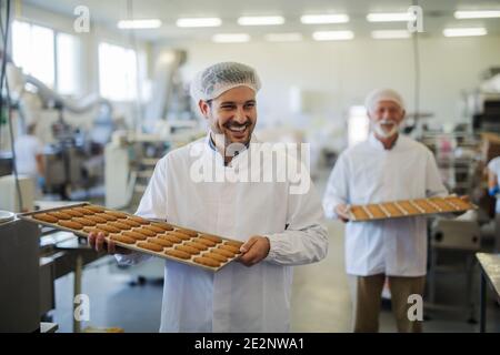 Photo de deux hommes souriants heureux dans des vêtements stériles portant des plateaux pleins de biscuits frais dans l'usine alimentaire. S'aider les uns les autres et regarder Banque D'Images