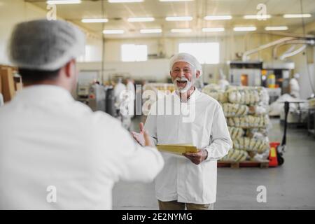 Image d'un homme âgé mûr et gai dans des vêtements stériles debout dans une usine alimentaire et être heureux de voir son collègue. Travail d'équipe et bon travail de collaboration Banque D'Images