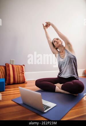 une femme est assise sur un tapis de yoga devant un ordinateur portable et s'étire Banque D'Images