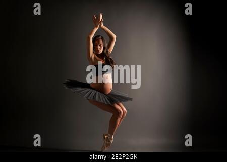 Jeune ballerine enceinte effectuant une posture classique de ballet avec le dessus noir et tutu en studio Banque D'Images