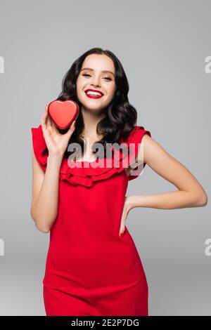 femme gaie en robe rouge tenant une boîte cadeau en forme de coeur pendant debout avec la main sur la hanche isolée sur le gris Banque D'Images