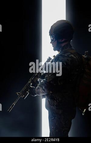 Photo d'un soldat dans un gilet blindé, un casque, des lunettes et un fusil, vue du profil de la rade sur fond ombré avec du brouillard. Banque D'Images