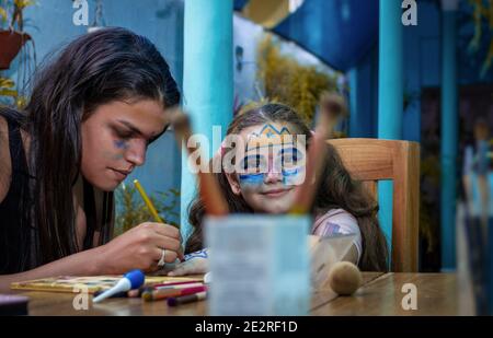 Fille avec des dessins sur son visage regardant droit devant pendant une jeune femme dessine un dessin sur la main de la fille Banque D'Images