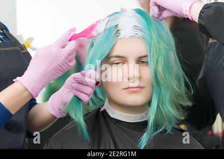 Processus de blanchiment des racines de cheveux dans salon de coiffure. Deux coiffeurs en gant de protection utilisent une brosse rose tout en appliquant de la peinture sur les cheveux émeraude du client Banque D'Images