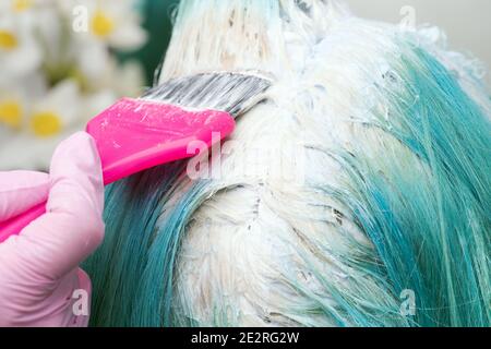 Gros plan du coiffeur dans un gant de protection à l'aide d'une brosse rose tout en appliquant de la peinture à la cliente féminine avec une couleur de cheveux émeraude, traitement blanchissant les cheveux Banque D'Images