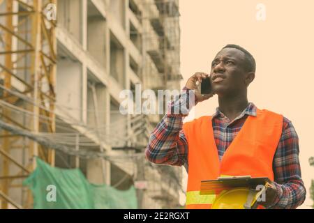 Young black African man construction worker talking on mobile phone en maintenant le casque de sécurité et presse-papiers au chantier Banque D'Images