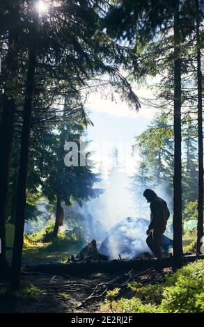 Gars en veste et capuche faisant feu et fumée s'écoule vers le haut. Tente parmi les sapins sous les rayons du soleil se brisant à travers les nuages par temps froid. Camping dans la forêt Banque D'Images