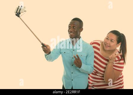 Joyeux jeune homme noir africain et jeune femme asiatique gras souriant tout en donnant le signe de la paix et en prenant des photos de selfie avec téléphone portable sur clé pour selfie Banque D'Images