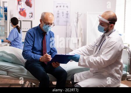 Patient âgé portant un masque facial lisant les documents administratifs au cours de la covid19. Spécialiste médical de la santé dans la salle de consultation clinique pendant la crise mondiale avec le coronavirus. Banque D'Images