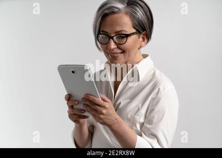 Portrait d'une femme âgée aux cheveux gris en chemise blanche et lunettes à l'aide d'une tablette numérique. Technologie par les personnes âgées qui naviguent sur Internet Banque D'Images