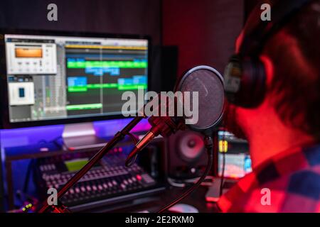 Le jeune homme blanc chante et enregistre une nouvelle piste dans le poste de travail de studio stéréo à l'aide d'un mixeur de chansons et d'un microphone. Banque D'Images