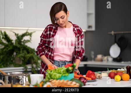 Couper des légumes frais jolie femme de ménage cuisine dîner portant une chemise à carreaux. Cuisiner avec passion jeune femme aux cheveux courts debout à la moderne Banque D'Images