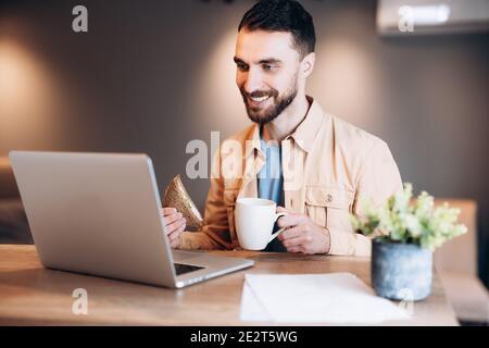 Joyeux travailleur indépendant travaillant à domicile. Jeune homme attrayant souriant, utilisant son revers moderne et parlant par videocall. Cuisine confortable en arrière-plan Banque D'Images