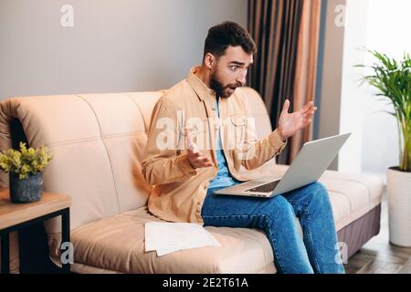 Le programmeur travaillant à domicile est choqué de lire le chat de travail. Jeune homme barbu dans des vêtements décontractés s'assoit sur un élégant canapé moderne avec un ordinateur portable sur ses genoux, son Banque D'Images