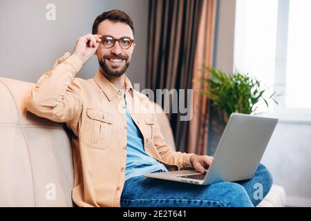 L'homme est satisfait du travail à distance. Un jeune homme en vêtements décontractés sourit en montrant ses dents et fixe ses lunettes tout en étant assis sur un canapé avec un ordinateur portable. Travail Banque D'Images