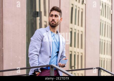 Jeune homme à barbe heureux tenant un smartphone tout en étant debout à l'extérieur et regarder loin Banque D'Images